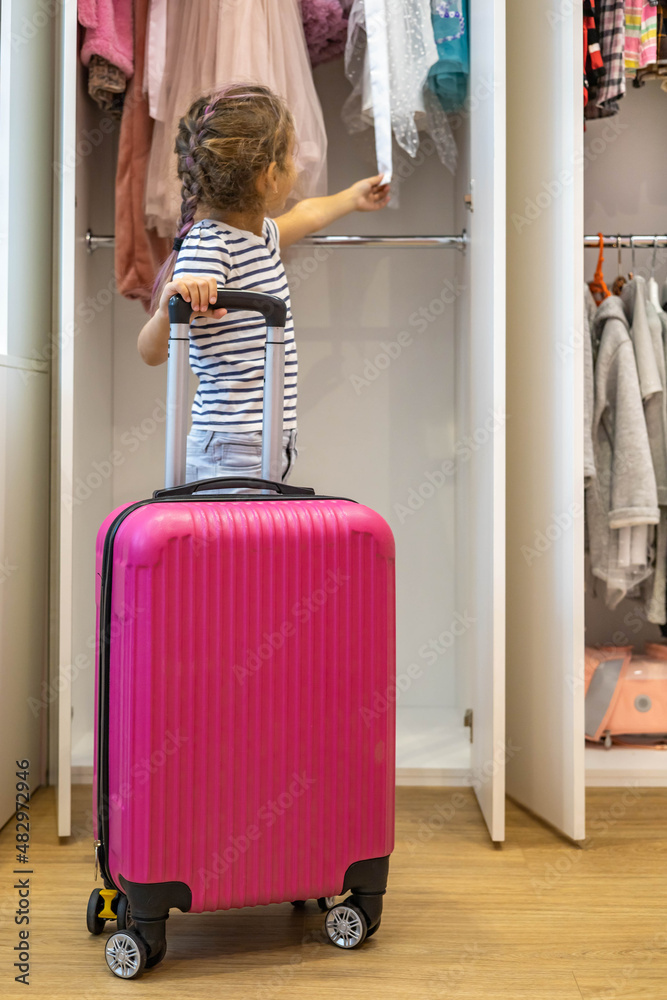 Little baby girl choosing clothes summer travel vacation with pink suitcase standing near cupboard Little baby girl choosing clothes summer travel vacation with pink suitcase standing near cupboard