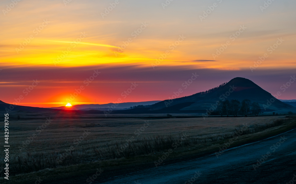 a landscape of a sunny sunrise, sunset among hills, high mountains and an agricultural field in the dark twilight