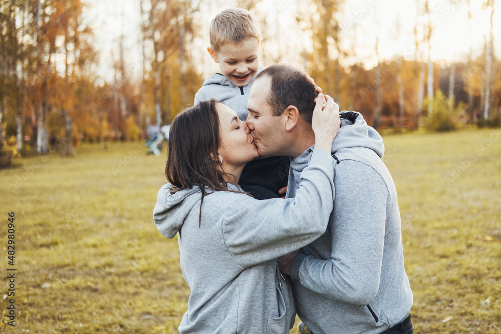 Mom and dad holding their son on their shoulders while they kiss and the child happily watches, spending happy time in the park. Portrait of parents kissing Mom and dad holding their son on their shoulders while they kiss and the child happily watches, spending happy time in the park. Portrait of parents kissing