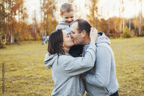 Mom and dad holding their son on their shoulders while they kiss and the child happily watches, spending happy time in the park. Portrait of parents kissing