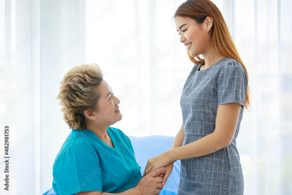 Obraz premium Old senior Asian happy healthy recovery mother patient in hospital uniform sitting smiling on bed while young woman daughter visiting standing holding hands with love and care in clinic wardroom