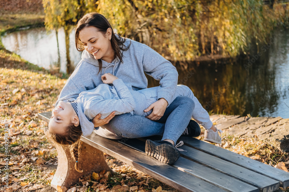 Fototapeta premium Mom sitting on the park bench holding her little girl lying on her arms laughing together. Family care. Happy family leisure. Smiling happy child.
