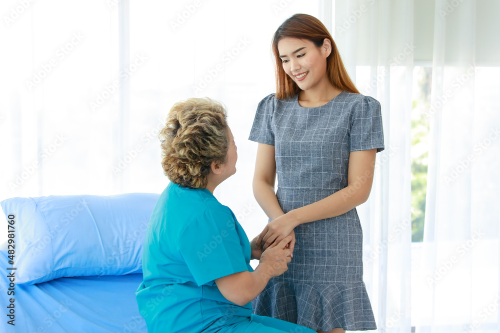 Old senior Asian happy healthy recovery mother patient in hospital uniform sitting smiling on bed while young woman daughter visiting standing holding hands with love and care in clinic wardroom Old senior Asian happy healthy recovery mother patient in hospital uniform sitting smiling on bed while young woman daughter visiting standing holding hands with love and care in clinic wardroom