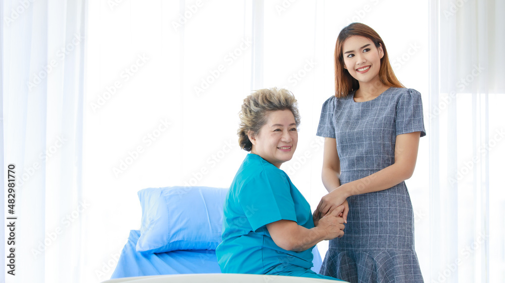 Old senior Asian happy healthy recovery mother patient in hospital uniform sitting smiling on bed while young woman daughter visiting standing holding hands with love and care in clinic wardroom Old senior Asian happy healthy recovery mother patient in hospital uniform sitting smiling on bed while young woman daughter visiting standing holding hands with love and care in clinic wardroom
