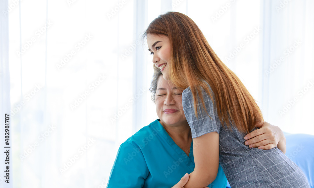 Old senior Asian happy healthy recovery mother patient in hospital uniform sitting smiling on bed while young woman daughter visiting standing holding hands with love and care in clinic wardroom Old senior Asian happy healthy recovery mother patient in hospital uniform sitting smiling on bed while young woman daughter visiting standing holding hands with love and care in clinic wardroom