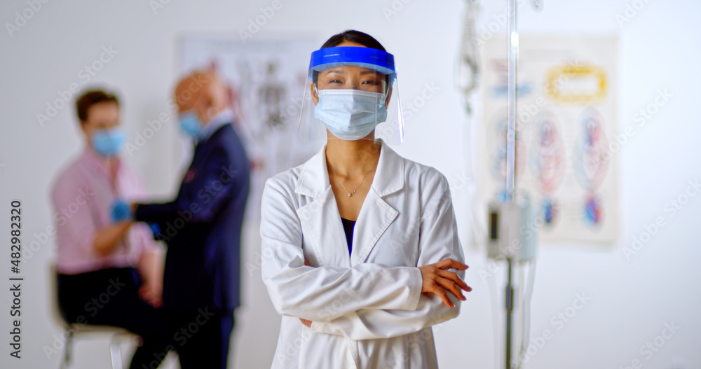 Young doctor with shield and face masks crosses arms looking towards camera smiling and confident as doctor helps patient in the background. Hopeful and optimistic, portrait