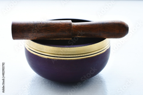 A close up image of a purple meditation singing bowl and wooden mallet on a white background. 