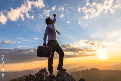 Successful business man, holding his business attache and standing on top of mountain goldden color sunset background, this photo conveys the message of success, competition and leadership concept.