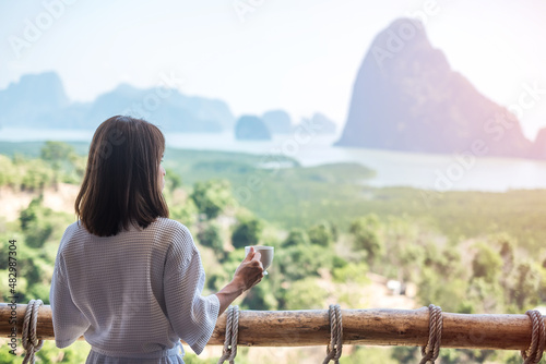 happy woman in bathrobe holding hot coffee cup or tea mug after waking up and enjoy Phang Nga bay view point, Tourist relaxing in tropical resort at Samet Nang She. Travel summer vacation concept