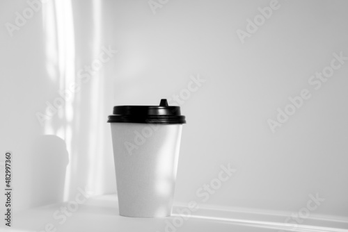 Mockup of paper cup with black plastic lid in rays of morning sunlight on white background.