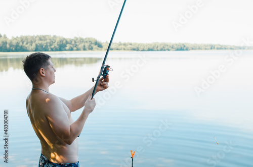 Young male fisherman with spinning rod standing on shore of lake on sunny summer day, outdoors. Side view of man fishing in nature. Hobby, sport and leisure concept