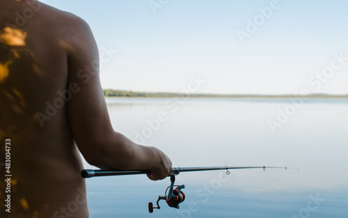Back view of male fisherman fishing on sunny summer day outdoors, copy space. Close-up of man holding spinning rod standing on shore in nature. Selective focus on fishing rod reel