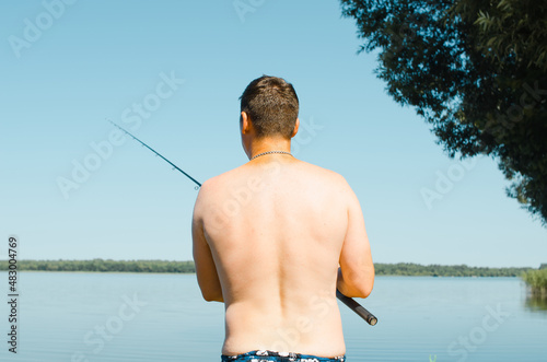Back view of male fisherman fishing on sunny summer day outdoors, waist length. Caucasian man holding spinning rod standing on shore of lake in nature. Sport and hobby, leisure and recreation concept