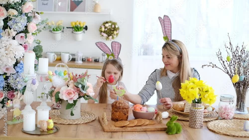 Cute little girl in bunny ears with her mother paint eggs while sitting at the table in the kitchen. Decorations for the celebration of Easter Cute little girl in bunny ears with her mother paint eggs while sitting at the table in the kitchen. Decorations for the celebration of Easter