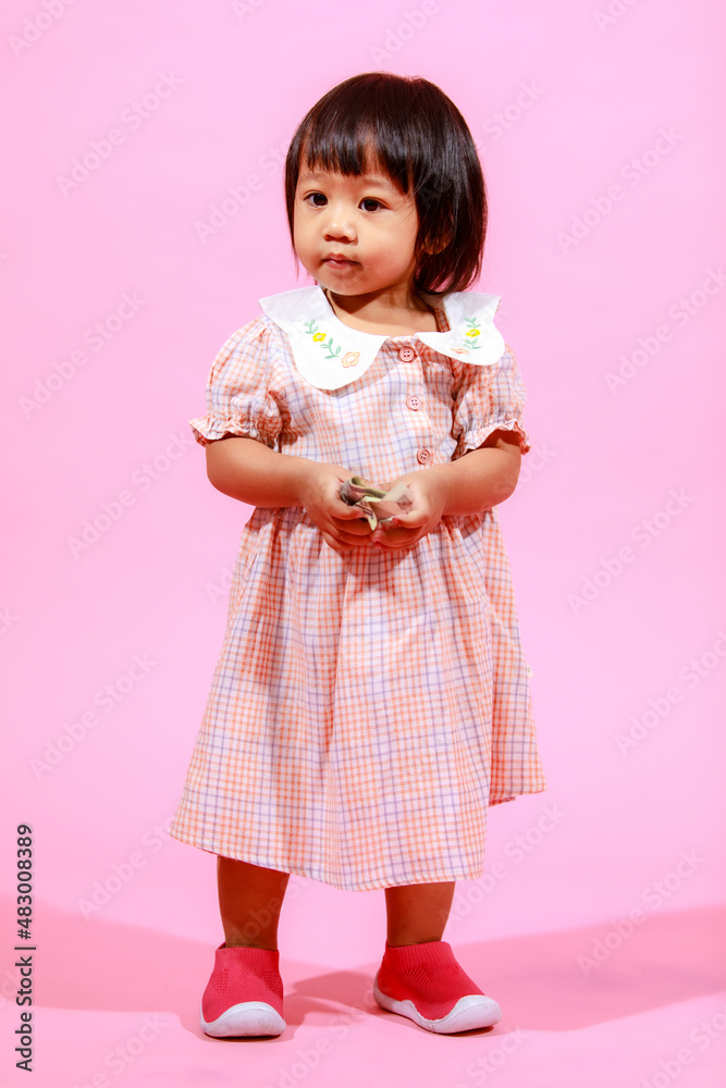 Portrait studio shot little cute Asian kindergarten preschooler girl daughter model in casual long dress and shoes standing look at camera holding hundred dollars banknote in hands on pink background Portrait studio shot little cute Asian kindergarten preschooler girl daughter model in casual long dress and shoes standing look at camera holding hundred dollars banknote in hands on pink background