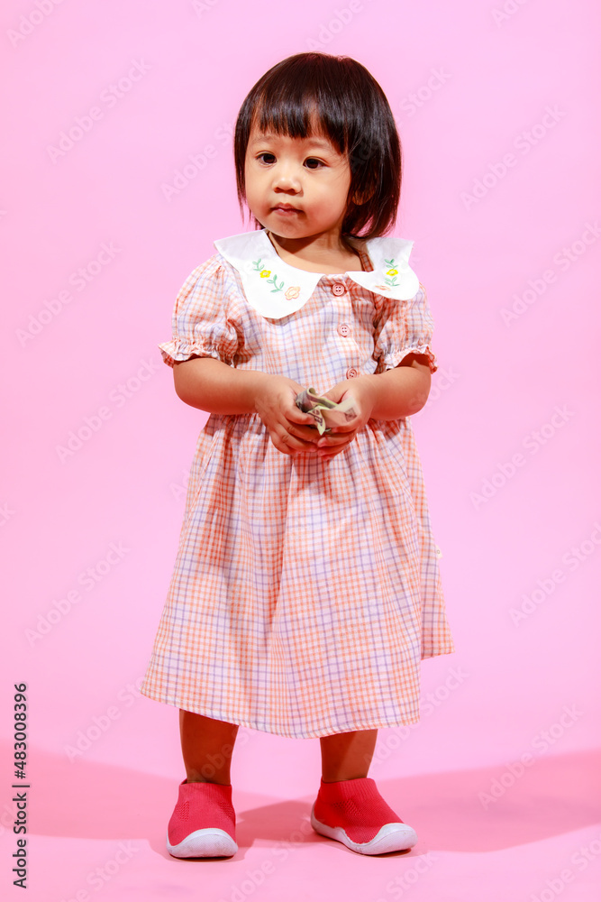 Portrait studio shot little cute Asian kindergarten preschooler girl daughter model in casual long dress and shoes standing look at camera holding hundred dollars banknote in hands on pink background Portrait studio shot little cute Asian kindergarten preschooler girl daughter model in casual long dress and shoes standing look at camera holding hundred dollars banknote in hands on pink background