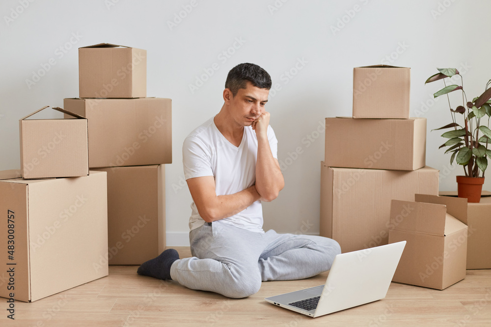 Sad bored man wearing white T-shirt sitting on the floor near cardboard boxes with personal pile during moving to new flat, looking at laptop screen with upset facial expression. Sad bored man wearing white T-shirt sitting on the floor near cardboard boxes with personal pile during moving to new flat, looking at laptop screen with upset facial expression.