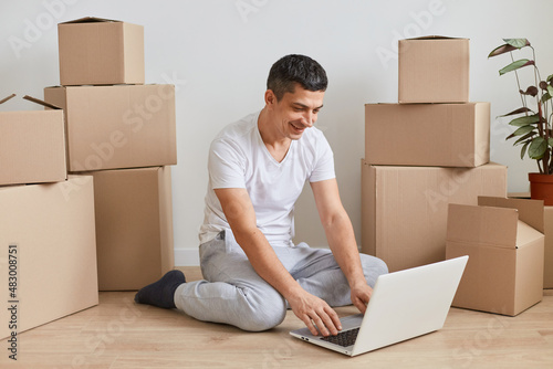 Portrait of young adult happy man wearing white T-shirt sitting on the floor near cardboard boxes with belongings, working on laptop with positive and happy facial expression.