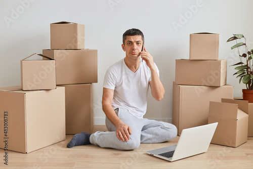 Image of sad upset man wearing white T-shirt sitting on the floor near cardboard boxes with personal pile, looking at camera with pout lips while talking phone, working on notebook.