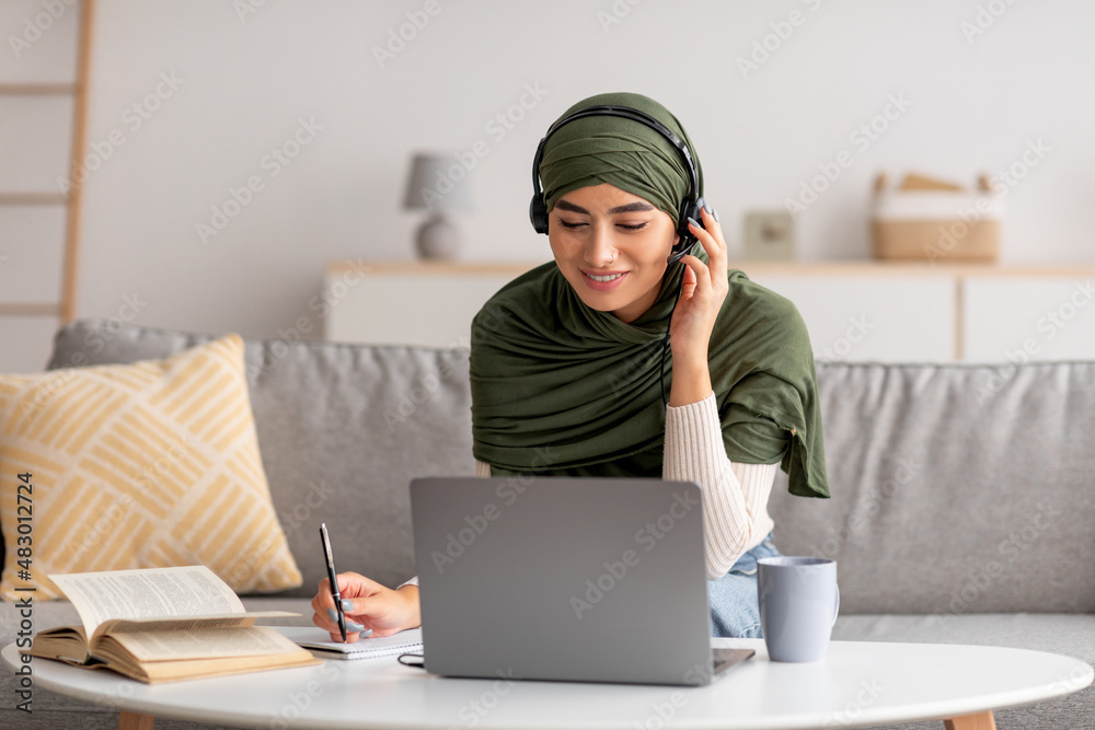 Millennial Arab woman in hijab using laptop, wearing headphones, taking notes during remote lesson at home Millennial Arab woman in hijab using laptop, wearing headphones, taking notes during remote lesson at home