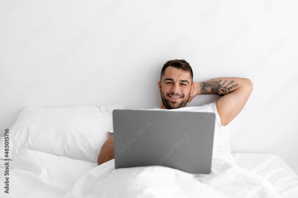 Satisfied relaxed millennial european muscular man lies on white bed and surfing in computer in bedroom Satisfied relaxed millennial european muscular man lies on white bed and surfing in computer in bedroom