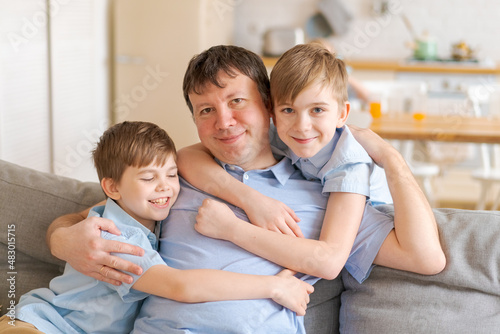 Happy caucasian father and little sons are sitting at home on sofa and having fun, playing hugging together. Excited dad and young children enjoy the family weekend with fun activities.