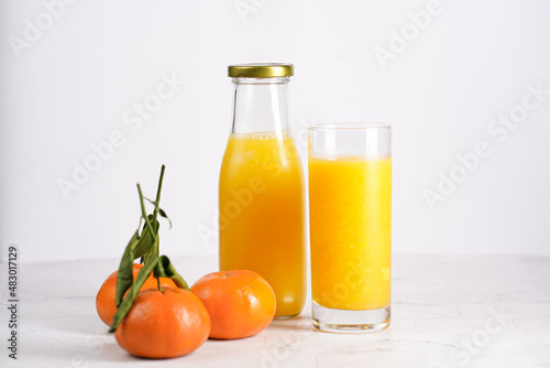 Tall glass with orange mango smoothie, several tangerines on white surface