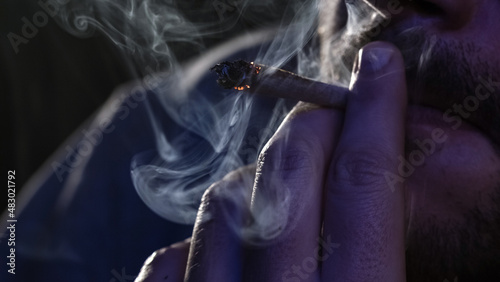 Male fingers with burning cannabis cigarette with smoke and cigarette glow under the blinking light, macro view.