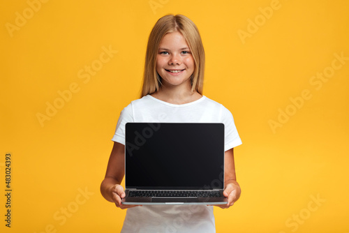 Cheerful adolescent blonde girl pupil show laptop with blank screen, isolated on yellow background