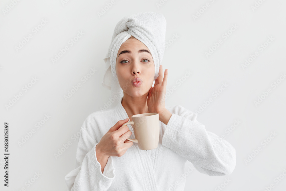 Smiling cute millennial caucasian blonde lady in bathrobe and towel on head, holds cup, drinks coffee Smiling cute millennial caucasian blonde lady in bathrobe and towel on head, holds cup, drinks coffee