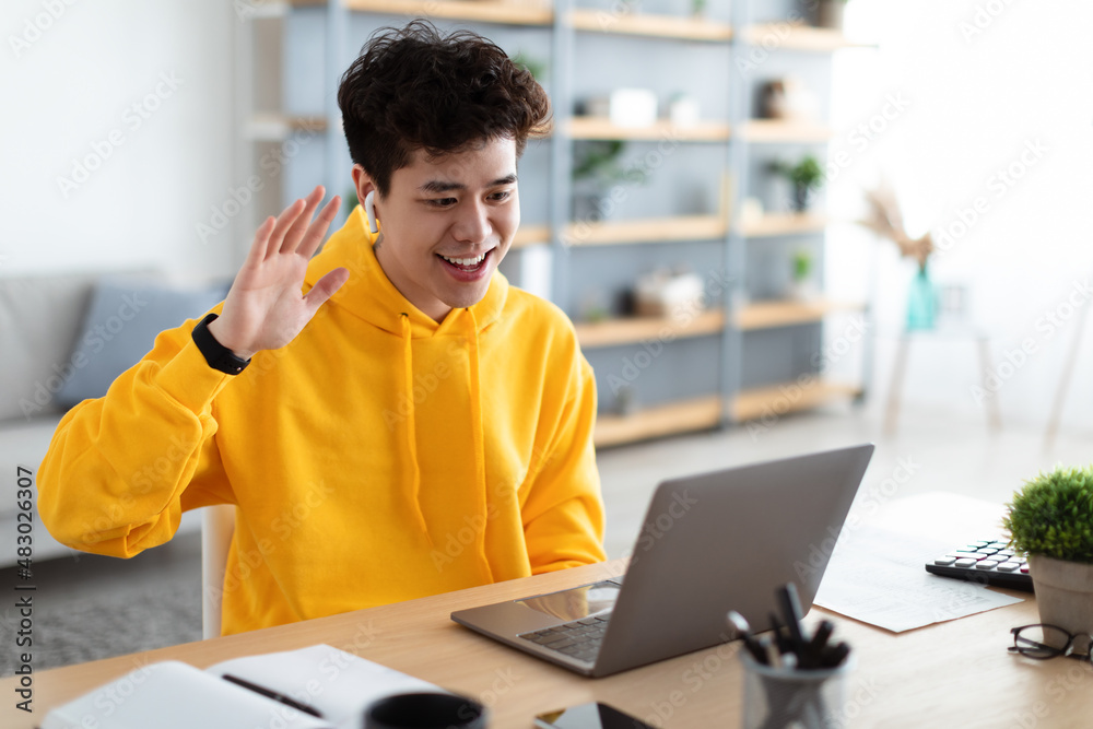 Fototapeta premium Smiling asian man working on laptop waving hand
