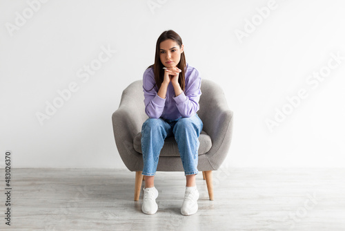 Full length of young woman sitting in armchair with dull or thoughtful face expression, having problem or depression