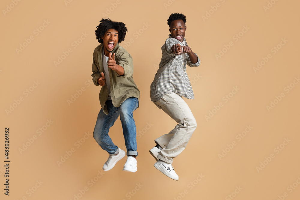 Two Cheerful Black Guys Pointing At Camera While Jumping Over Beige Background Two Cheerful Black Guys Pointing At Camera While Jumping Over Beige Background