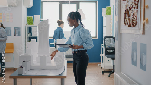 Engineer working on blueprints design with digital tablet and building model at work. Woman architect using device to plan construction structure and layout. Architectural project