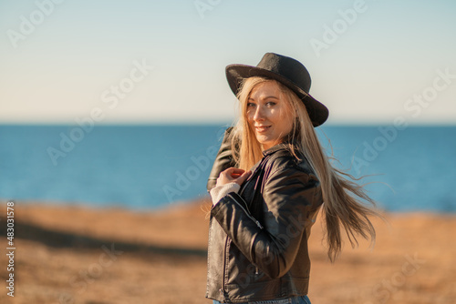A blonde in a stylish black leather jacket walks along the seashore. © svetograph
