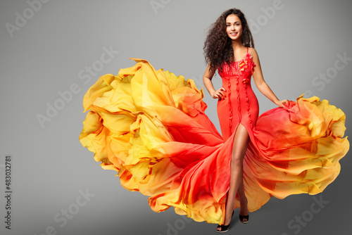 Woman Dancing in Colorful Yellow Orange Flying Dress. Happy Girl Model in Creative Bright Gown over Gray Background. Curly Latin Salsa Dancer