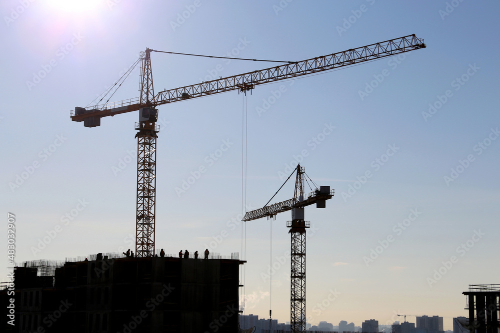 Silhouette of construction cranes and workers on unfinished residential building against sunshine. Housing construction, apartment block in city