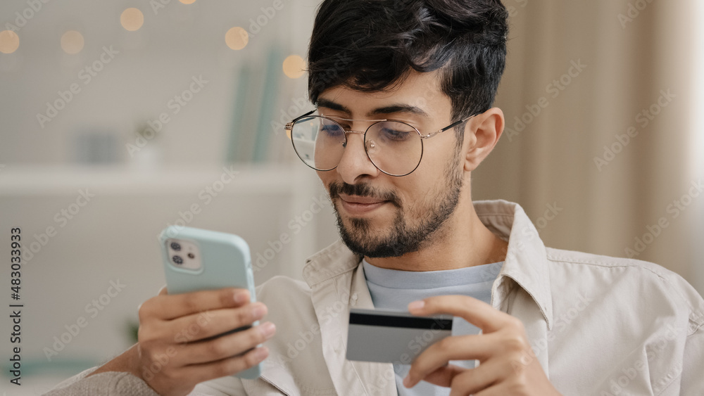 Close-up bearded arabic business man in glasses with credit card and phone makes purchase online shopping enters bank number online transaction uses mobile financial application booking order buying Close-up bearded arabic business man in glasses with credit card and phone makes purchase online shopping enters bank number online transaction uses mobile financial application booking order buying