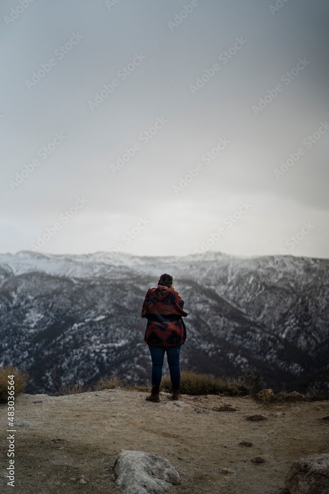Chica con poncho colorido tomando fotos y sonriendo al borde de la ladera con la sierra nevada de fondo