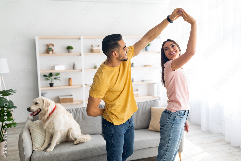 Romantic young diverse couple dancing at home, their cute dog sitting on couch nearby, copy space