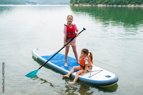 Girls padding on stand up paddle boarding on lake district. Children in swim life vest learning paddleboarding on SUP board. Family active leisure and local getaway with kids concept photo