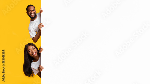 Black couple peeking out blank white advertising billboard at studio