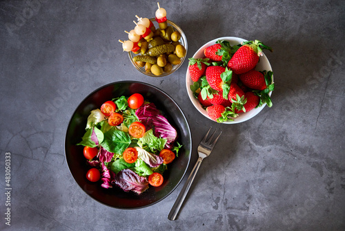 A salad, a bowl of chips, a bowl of Spanish brochettes and another bowl of strawberries on a black slate background.