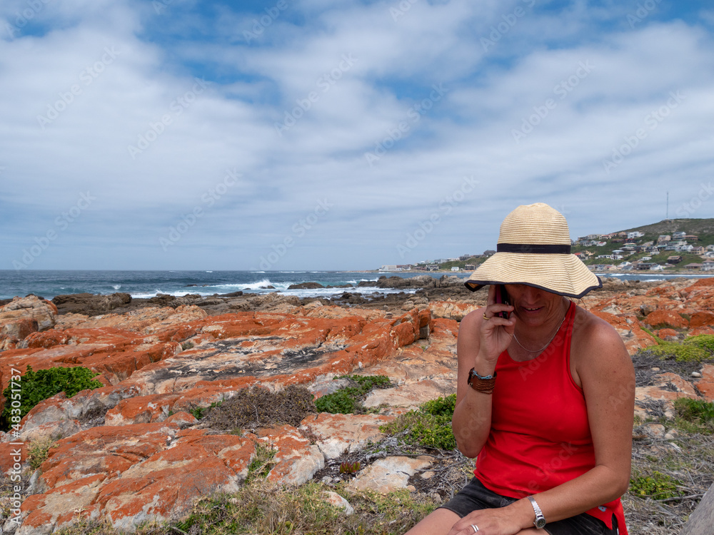 A caucasian woman chatting on her cellphone while she is sitting on rocks next to the Atlantic Ocean. Location: Jongensfontein, Western Cape