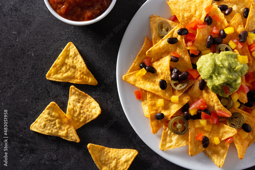 Mexican nachos tortilla chips with black beans, guacamole, tomato and jalapeno on black background Mexican nachos tortilla chips with black beans, guacamole, tomato and jalapeno on black background
