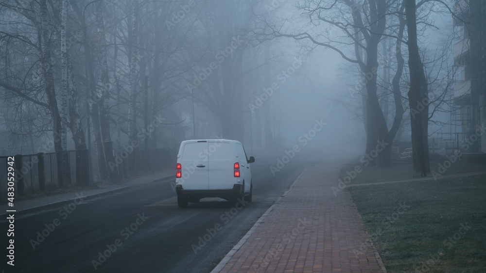  White commercial vehicle driving through the foggy and mysterious city road during a cold autumn morning, motion-blurred car