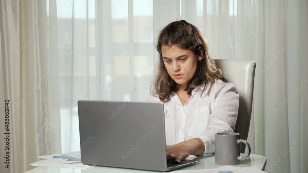 Disabled woman freelancer having cerebral palsy takes cup with problem to drink tea working from home on computer sitting at table against window. Disabled woman freelancer having cerebral palsy takes cup with problem to drink tea working from home on computer sitting at table against window.