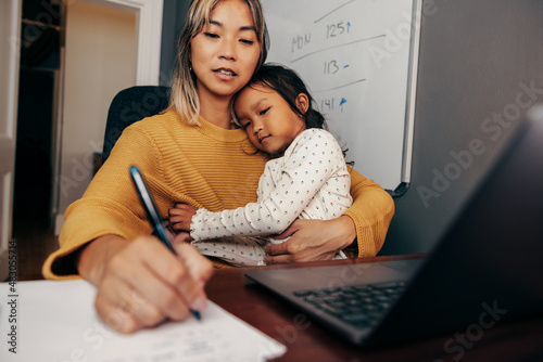 Young working mom writing notes while embracing her daughter
