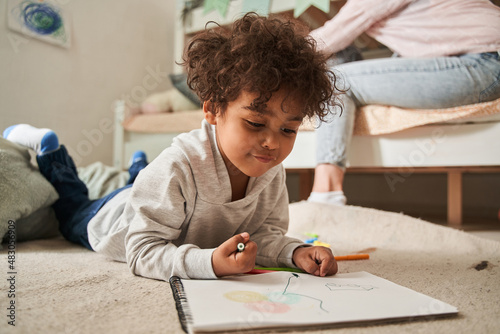 Little multiracial boy laying at the floor and drawing something with pencils at the paper