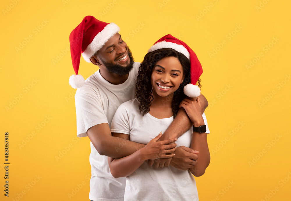 Loving african american couple in Santa hats hugging on yellow studio background, celebrating New Year or Christmas Loving african american couple in Santa hats hugging on yellow studio background, celebrating New Year or Christmas